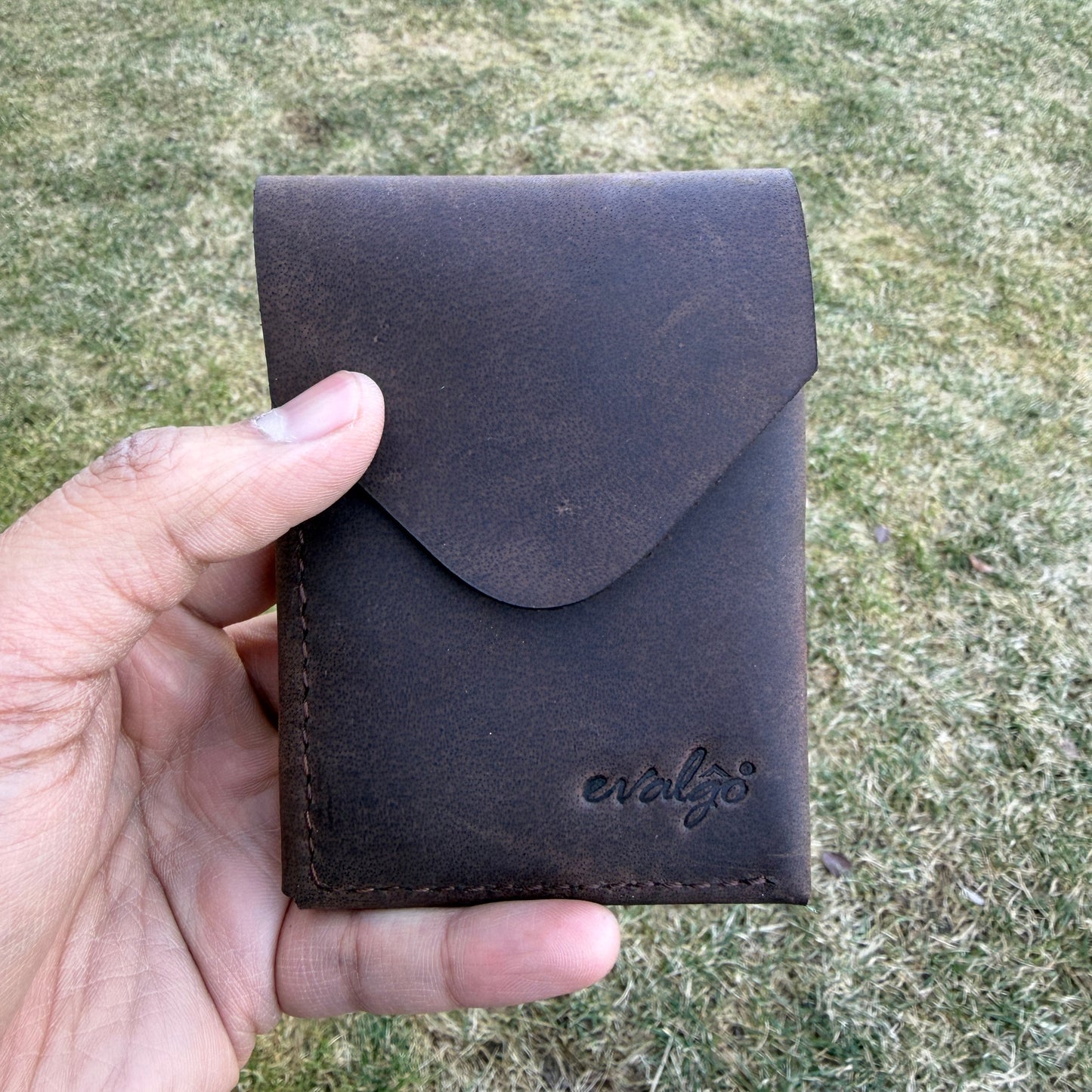 Hand holding a dark brown leather wallet against a grassy background