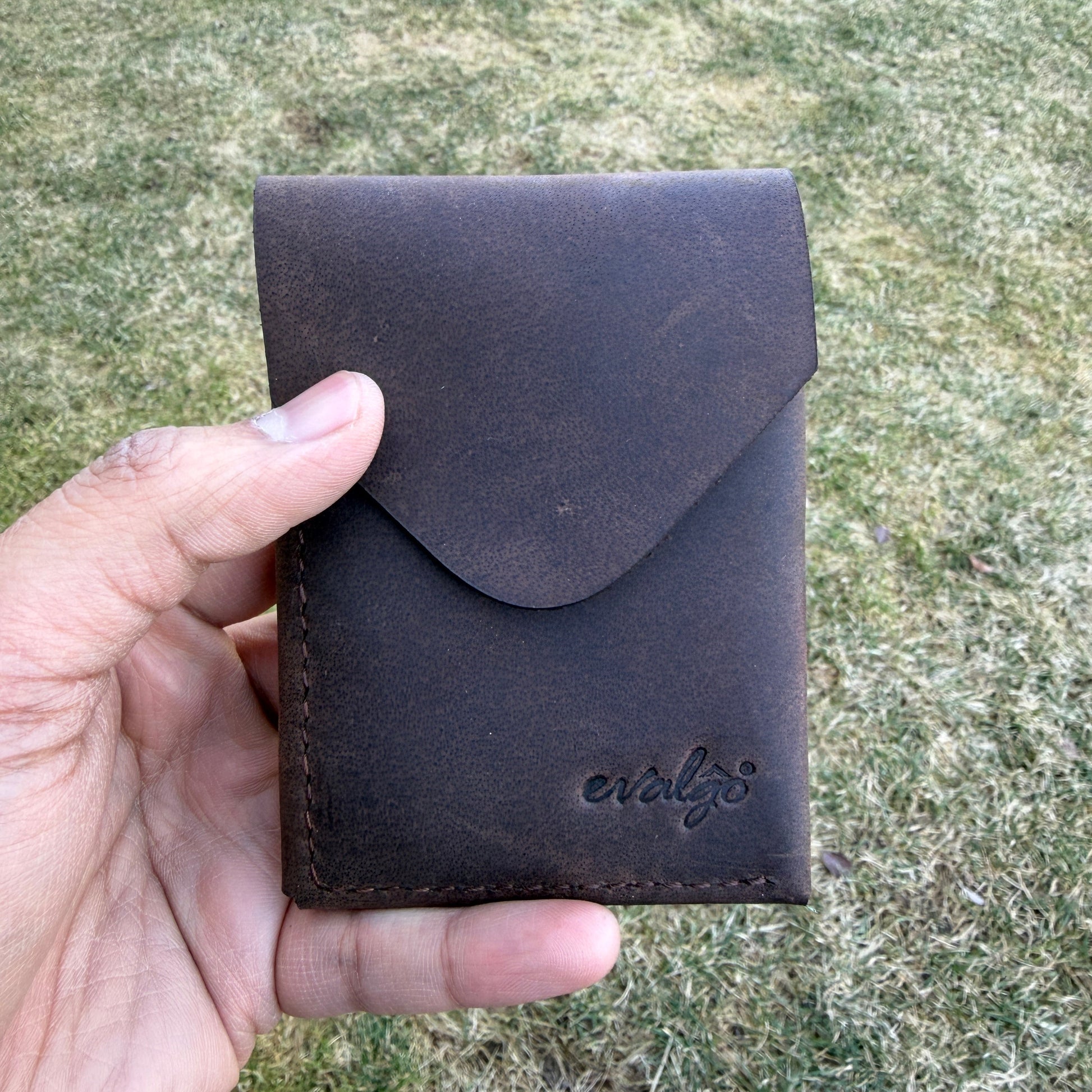 Hand holding a dark brown leather wallet against a grassy background