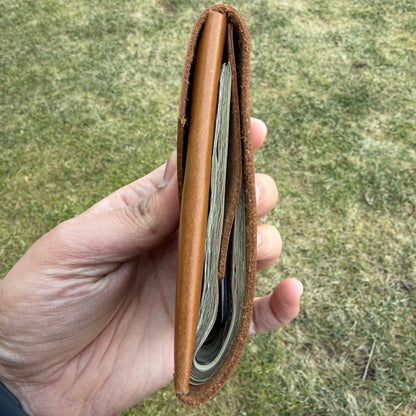 Hand holding a brown leather wallet with money against a grassy background