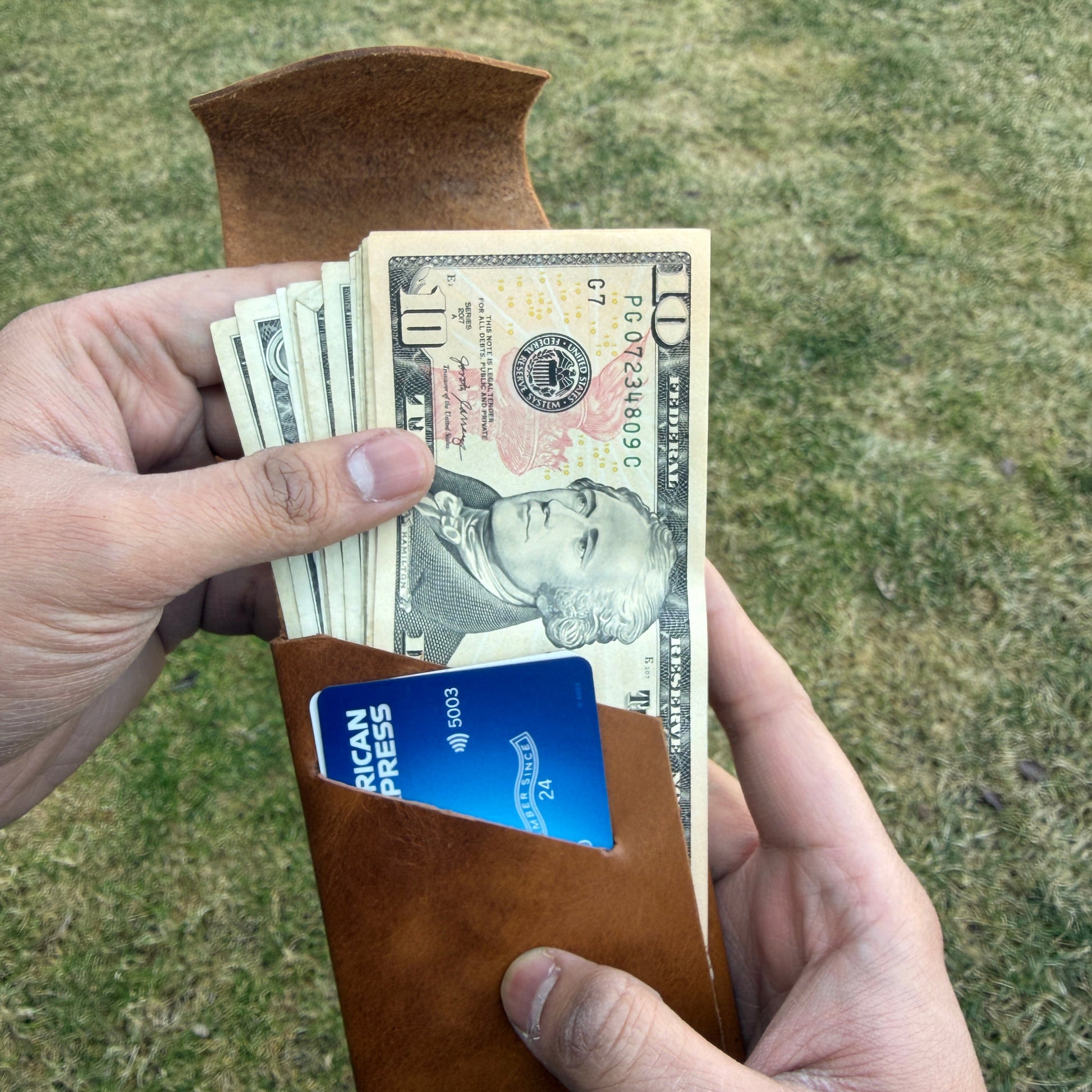 Person holding a brown leather wallet with money and a card against a grassy background